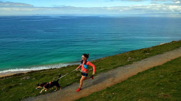 A woman wearing a running vest with her dog on a lead runs along a coast path with sea in the distance